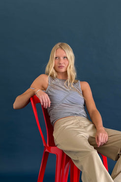 A blonde woman sits on a red plastic chair against a dark blue studio background, wearing a gray sleeveless ruched top and taupe pants.