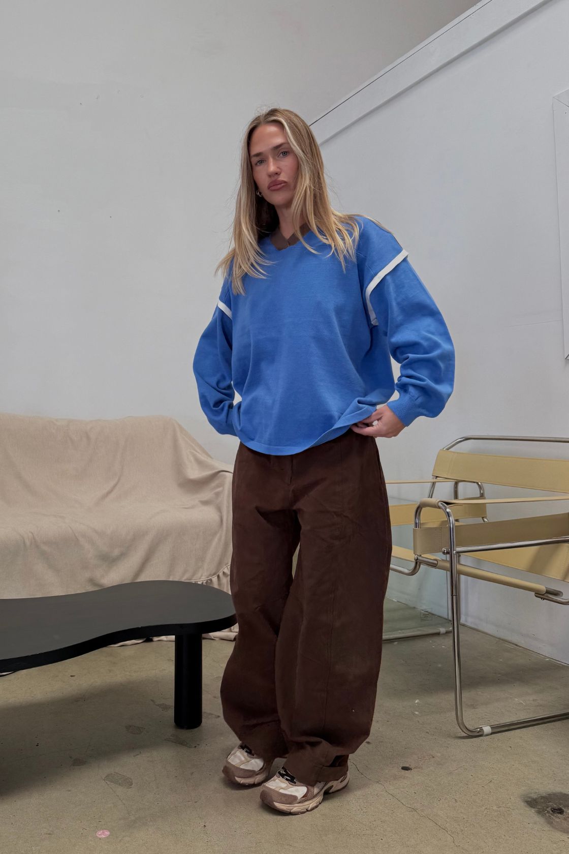 Lifestyle image of a model wearing the Eames Pants in espresso featuring a soft velvet-style cotton finish, relaxed straight-leg fit and clean waistband, styled with a bright blue oversized sweatshirt and neutral sneakers in a minimal studio setting