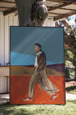 A large framed painting leaning outdoors against a tree trunk on grass. The artwork depicts a person walking across a surreal landscape with a dark blue sky, orange-red ground, and small horse figures.