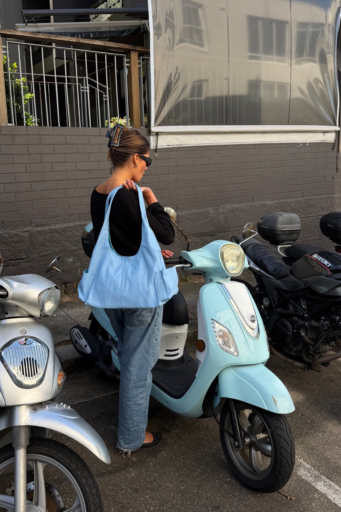 A woman stands next to a light blue scooter in an outdoor parking area, wearing a black top, blue jeans, and carrying a large light blue leather bag.
