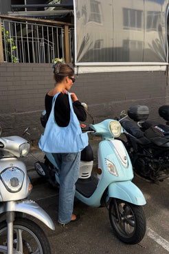 A woman stands next to a light blue scooter in an outdoor parking area, wearing a black top, blue jeans, and carrying a large light blue leather bag.