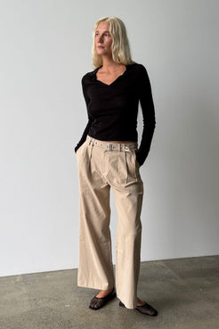 Model wearing the Soraya pant in Taupe with front pleats and a matching belt, styled with a fitted black long sleeve top and black woven flats.