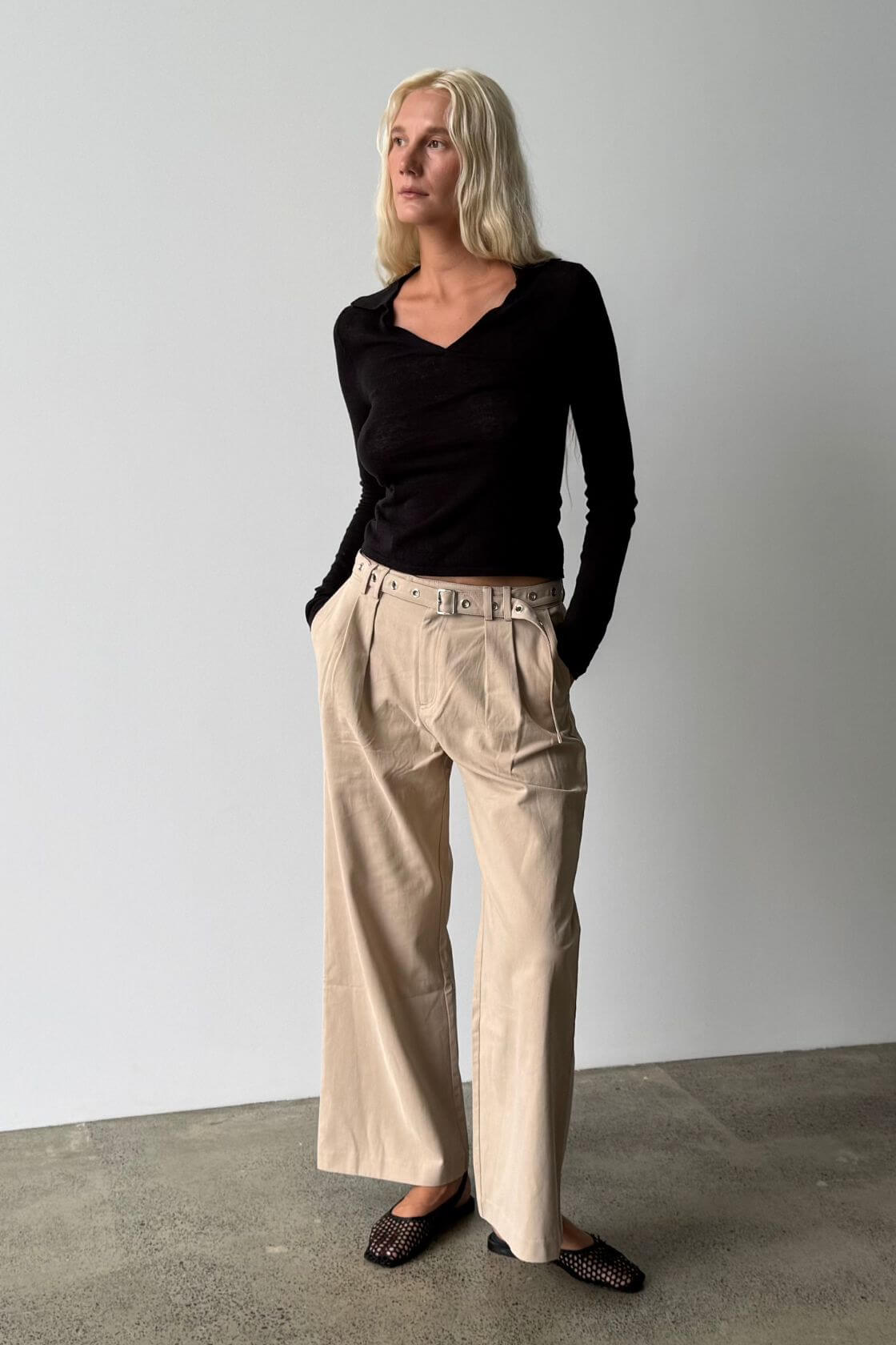 Model wearing the Soraya pant in Taupe with front pleats and a matching belt, styled with a fitted black long sleeve top and black woven flats.