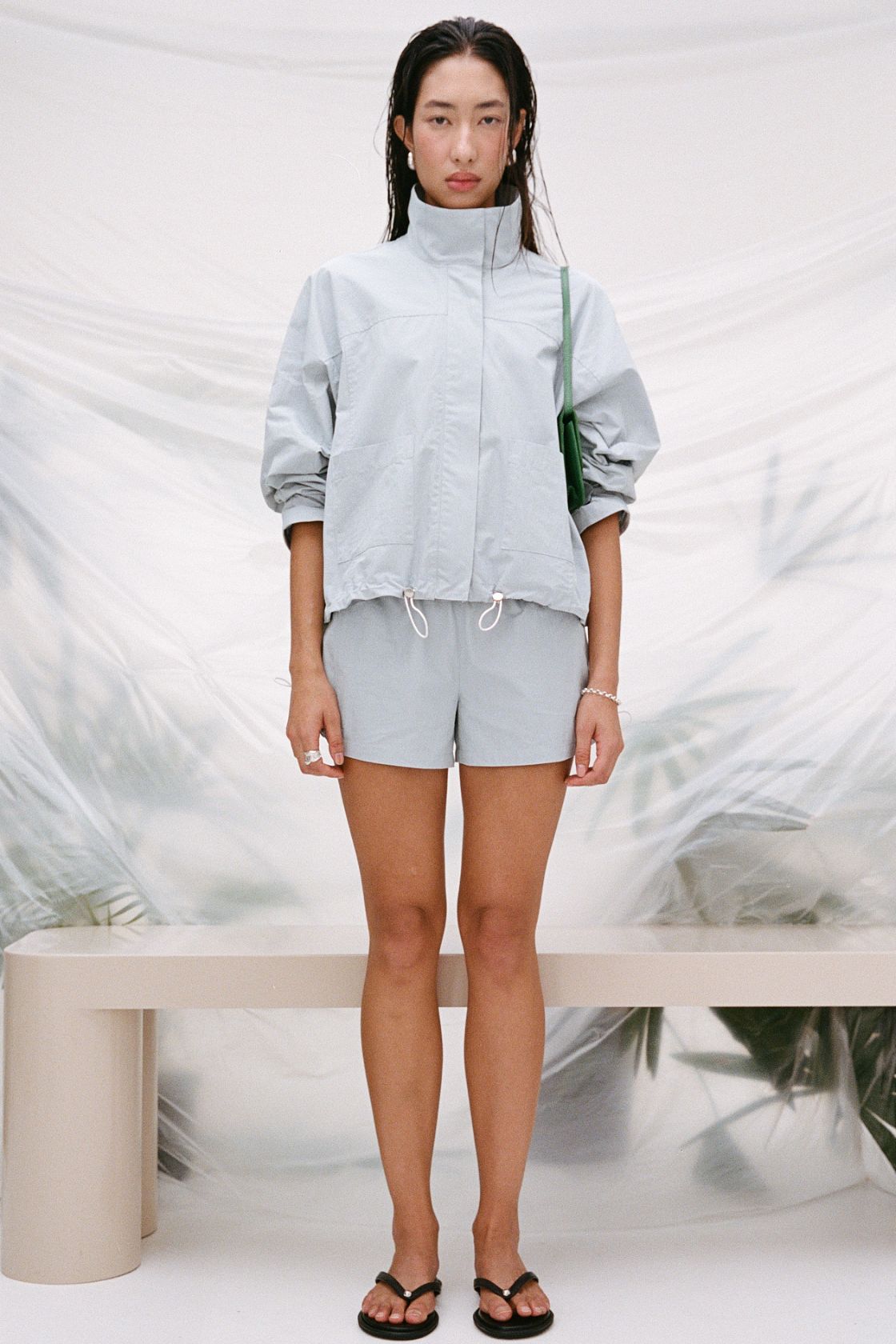 A model stands in a studio wearing the Addie jacket in light blue with matching light blue shorts, with a green shoulder bag and black flip-flops. The background features a draped white fabric backdrop with a beige bench.