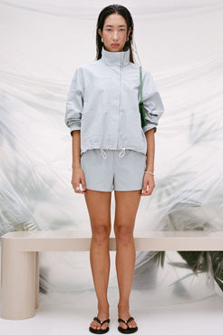 A model stands in a studio wearing the Addie jacket in light blue with matching light blue shorts, with a green shoulder bag and black flip-flops. The background features a draped white fabric backdrop with a beige bench.