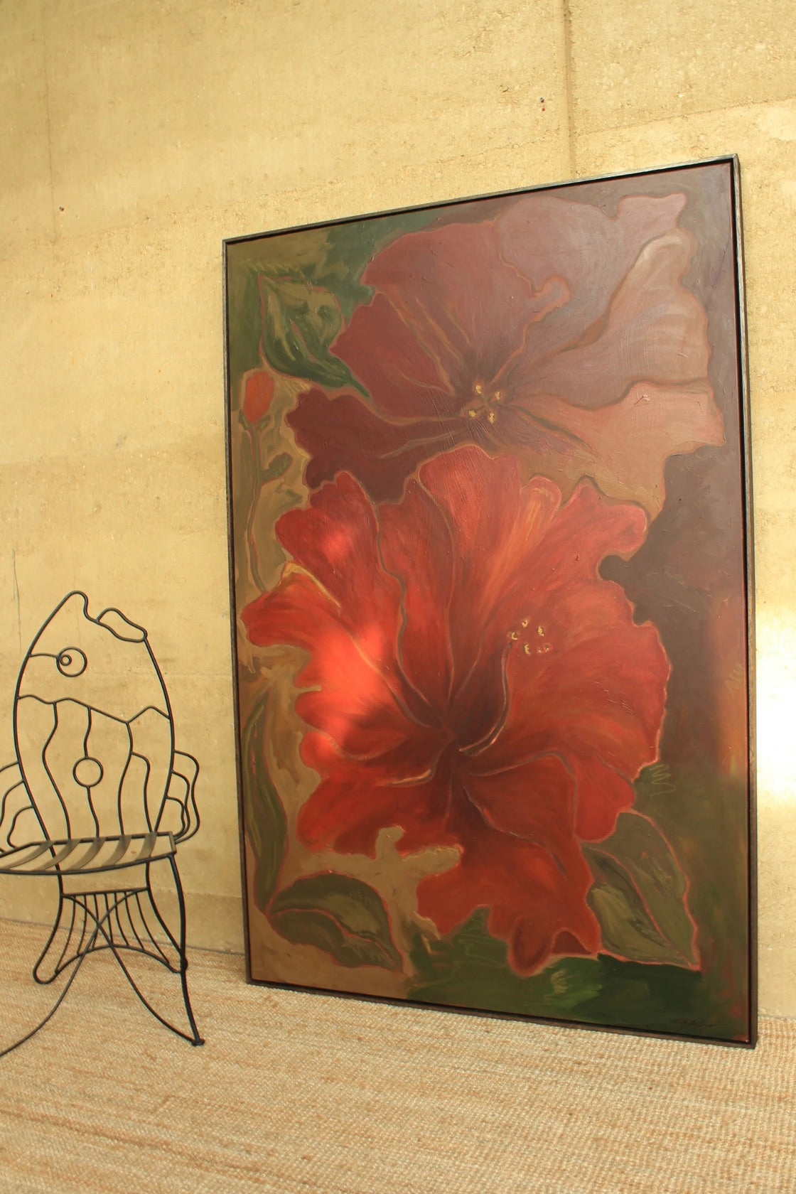 A framed painting of two large red and burgundy flowers with green leaves, leaning against a beige wall next to a black wire fish-shaped chair.