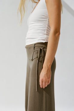 A side-profile view of a model wearing a white ruched tank top and high-waisted, wide-leg olive knit trousers with a side tie detail.