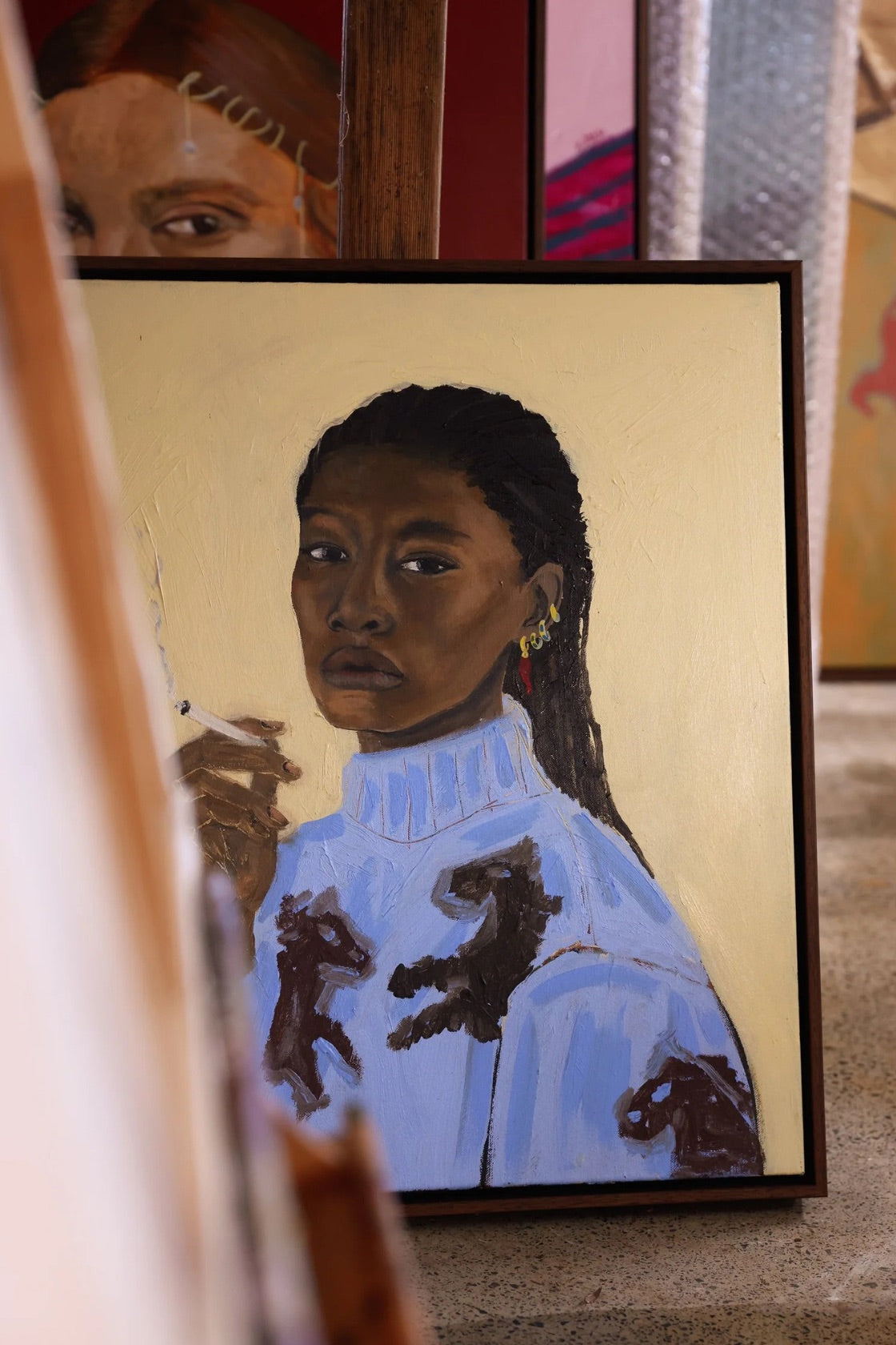 A framed portrait painting of a Black woman with braided hair, wearing a light blue turtleneck sweater with dark brown abstract marks, displayed on an easel in an art studio setting.