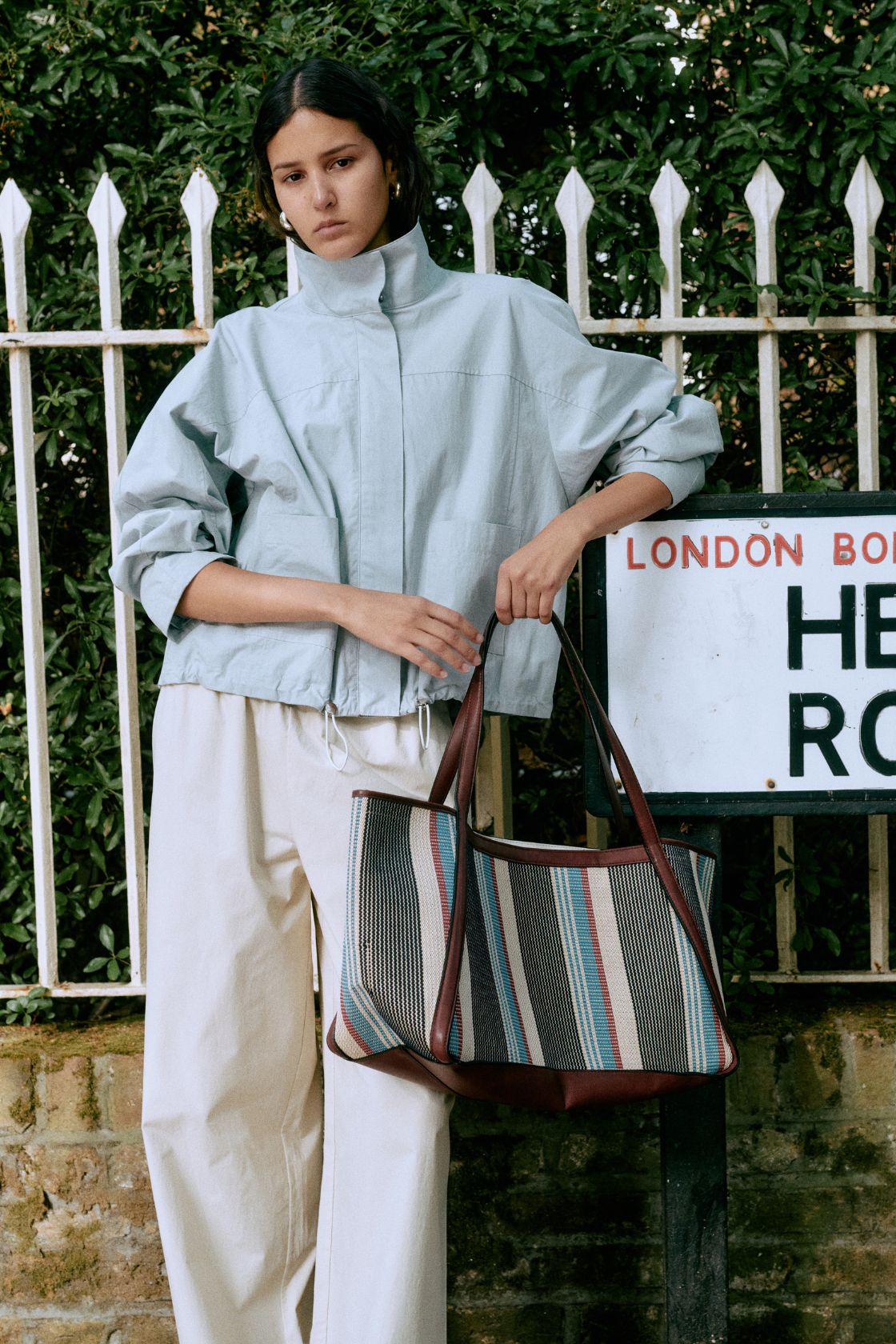 Model wearing the Addie jacket in pale blue with cream pants and brown striped bag against a white fence