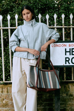 Model wearing the Addie jacket in pale blue with cream pants and brown striped bag against a white fence