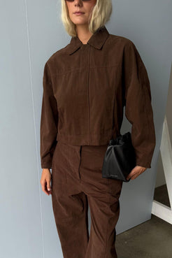 Model wearing an espresso cotton jacket with a concealed front closure and matching barrel-leg pants, styled with a black pouch bag for a tonal utilitarian look.