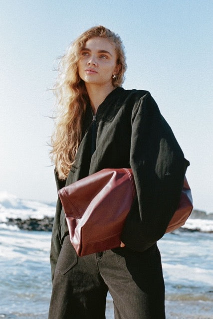 Lifestyle image of the Wren black bomber jacket worn outdoors by the beach with a burgundy leather bag