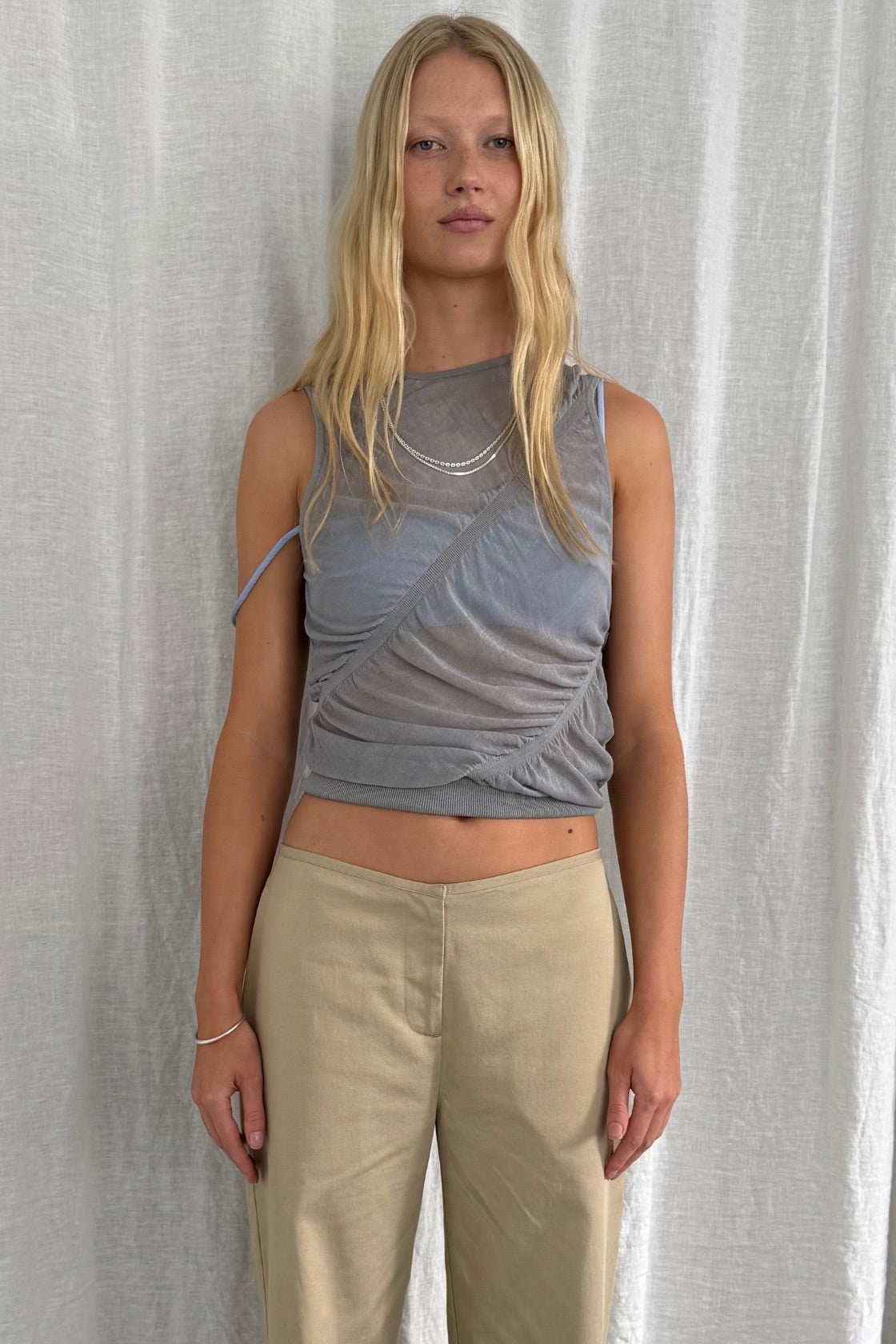 A blonde model stands against a white curtain backdrop wearing a ruched, semi-sheer gray sleeveless crop top with a mesh overlay and beige wide-leg pants.