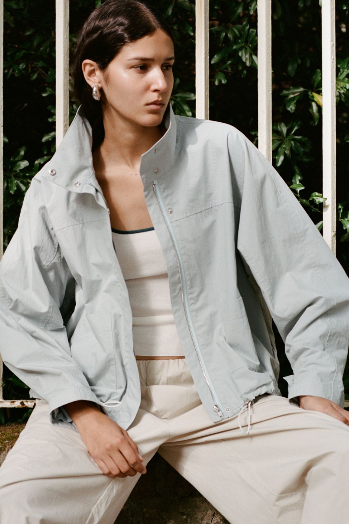 A woman wearing the Addie jacket in pale blue over a cream ribbed tank top and beige drawstring trousers, sitting outdoors in front of a white fence with green foliage behind her.