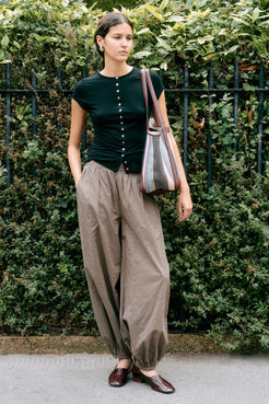 Model wearing Robbie top in black with Elin balloon pants in chocolate check against a background with green foliage. 