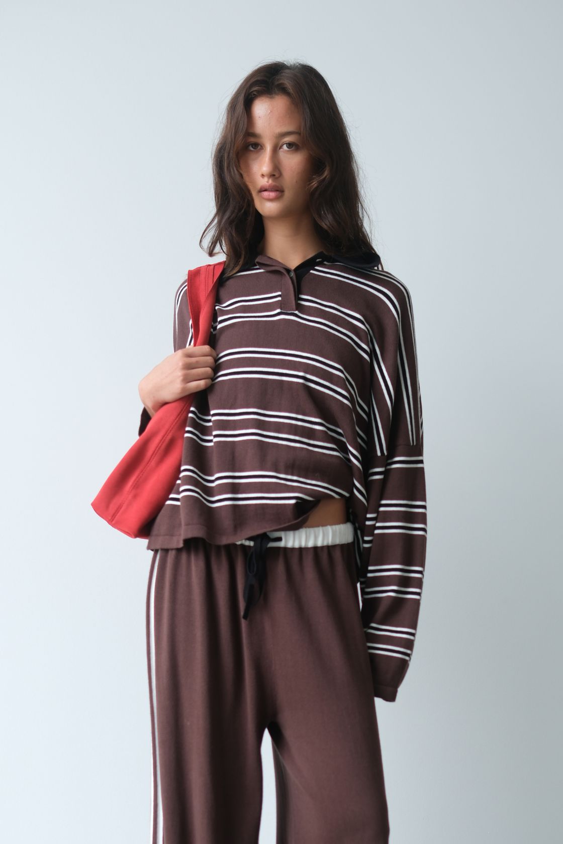 Model wearing the Rayne Jersey in Espresso, Cream & Black Stripe, featuring a relaxed collared silhouette with horizontal stripe detailing, styled with matching wide-leg pants and a red shoulder bag against a soft grey backdrop.