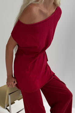 A woman wearing a chilli off-the-shoulder knit top and matching wide-leg pants, seated on a light-colored chair against a plain light gray background.