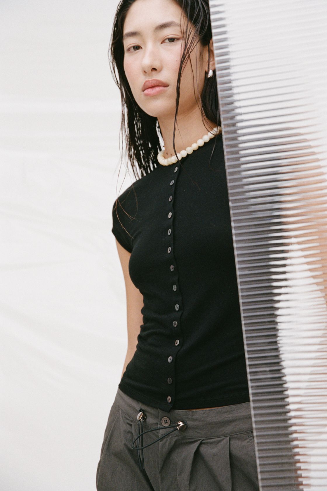 A woman wearing a fitted black short-sleeve top with a button front and gray pants, standing beside a white slatted screen against a light studio background.