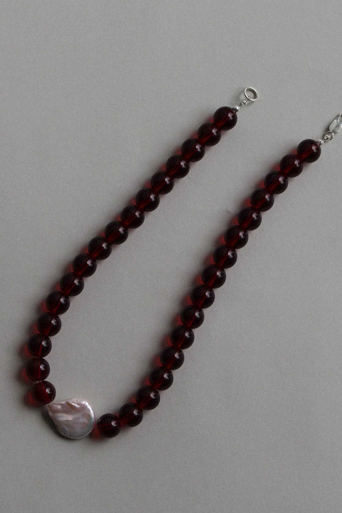 Red garnet beaded necklace with freshwater pearl and silver hardware, shown in a soft curve on a neutral grey surface.