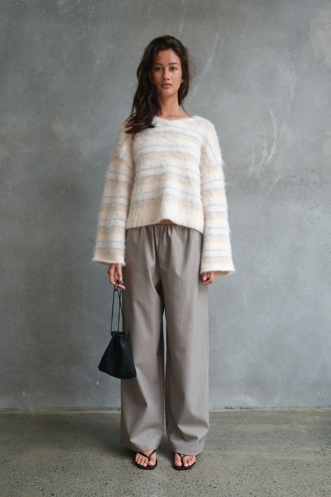 Model wearing the Lucca Sweater in Taupe & Blue Stripe, featuring a soft fluffy knit with subtle blue stripe detailing, styled with relaxed taupe wide-leg trousers, black flip flops and a black drawstring pouch bag against a textured concrete backdrop.