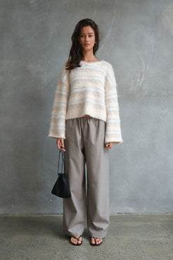 Model wearing the Lucca Sweater in Taupe & Blue Stripe, featuring a soft fluffy knit with subtle blue stripe detailing, styled with relaxed taupe wide-leg trousers, black flip flops and a black drawstring pouch bag against a textured concrete backdrop.