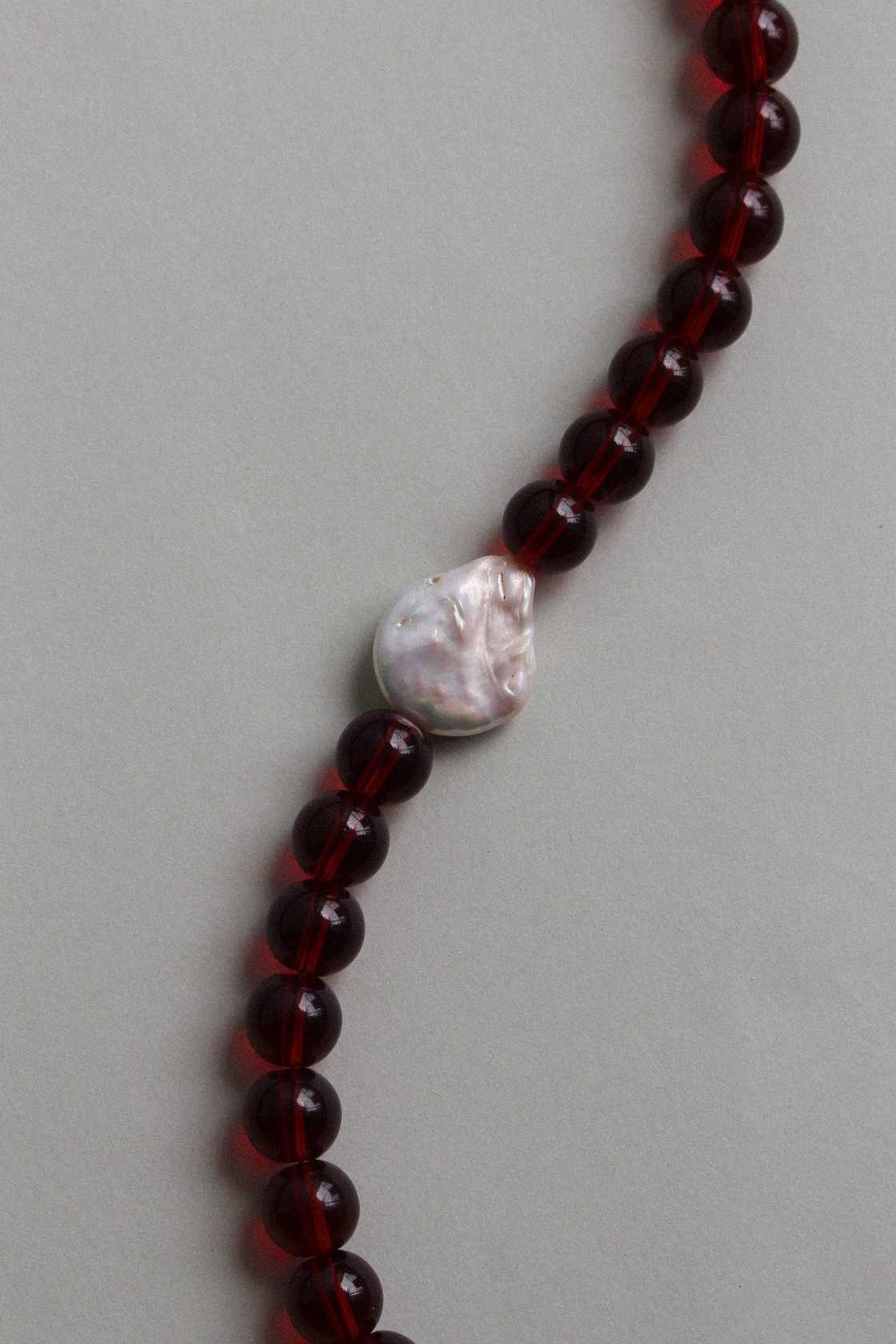 Close-up of polished red garnet beads and an irregular freshwater pearl on a minimal beaded necklace.