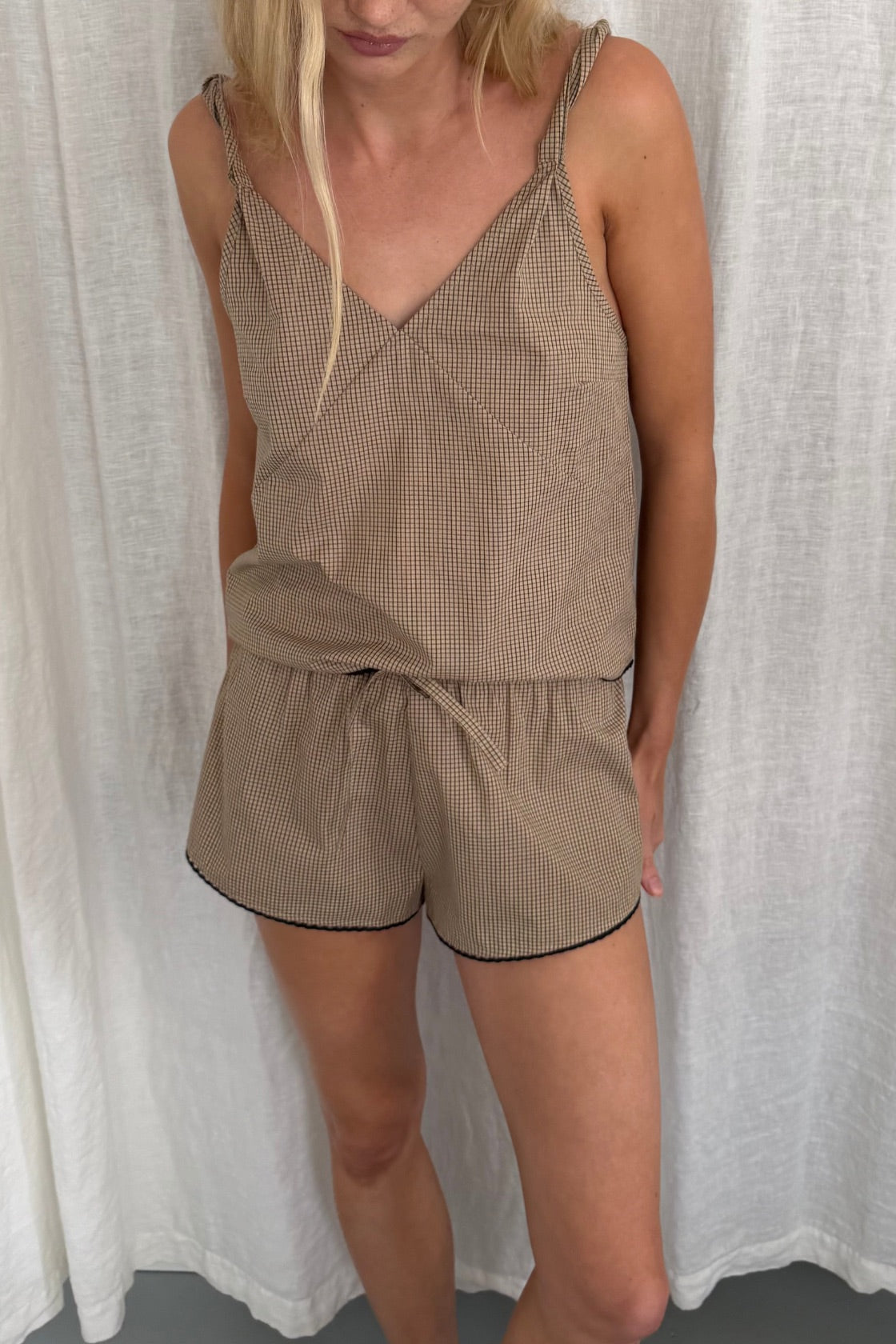 A woman wearing a taupe micro-checkered two-piece set consisting of a camisole top with thin straps and matching shorts with black piping, standing in front of a white curtain.