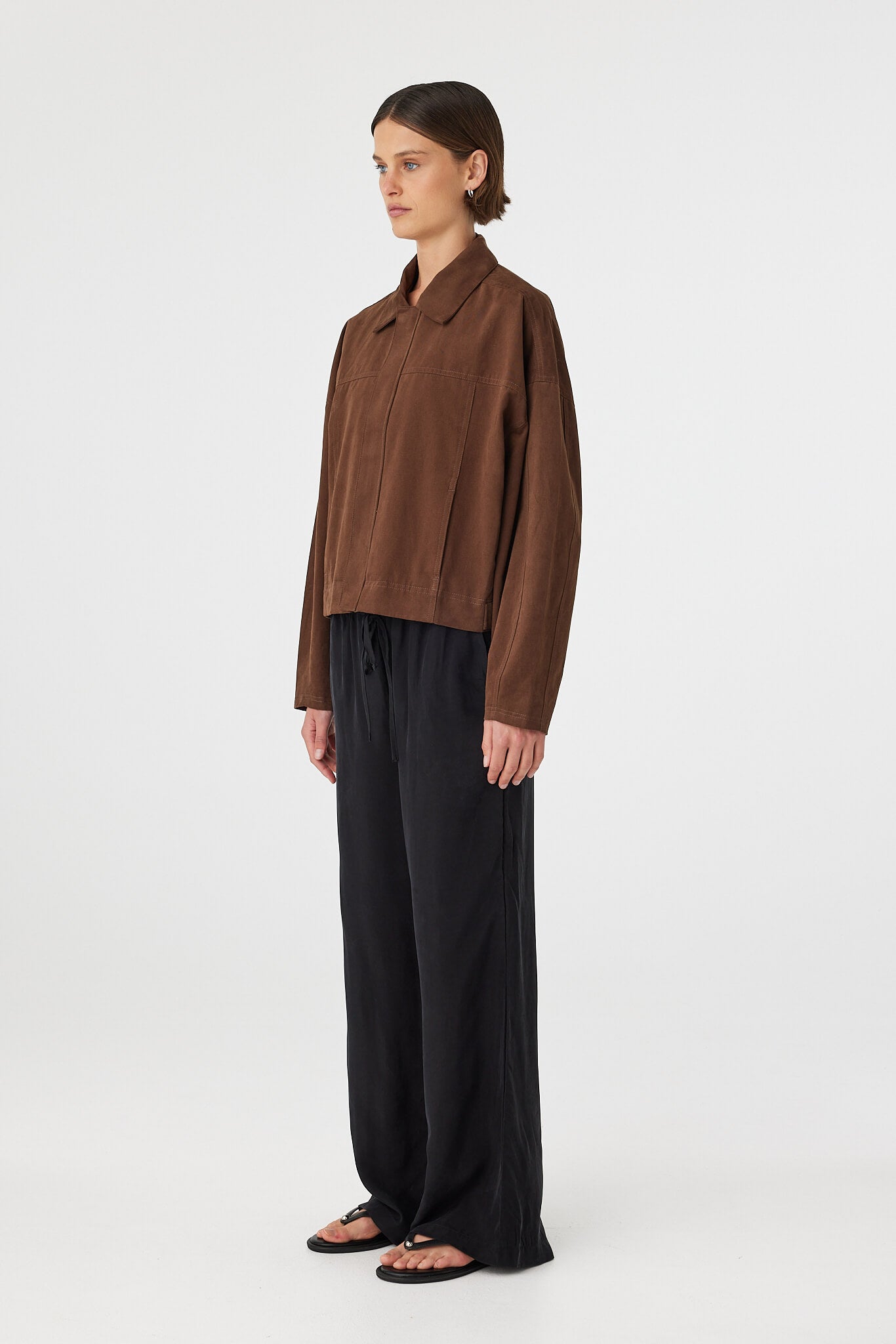 Eames jacket in espresso shown in side profile, highlighting the structured shape, dropped shoulders and cropped length, styled with black pants and sandals
