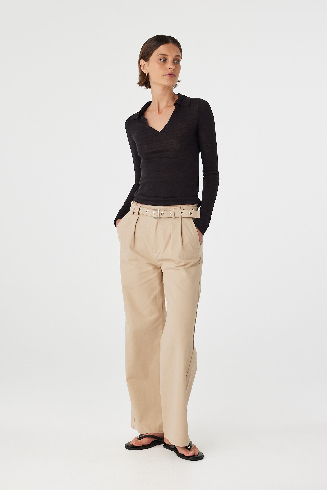Mae longsleeve top in black worn at an angle with hands in pockets, showcasing the fitted silhouette, collar neckline and soft knit texture, styled with beige pants and sandals