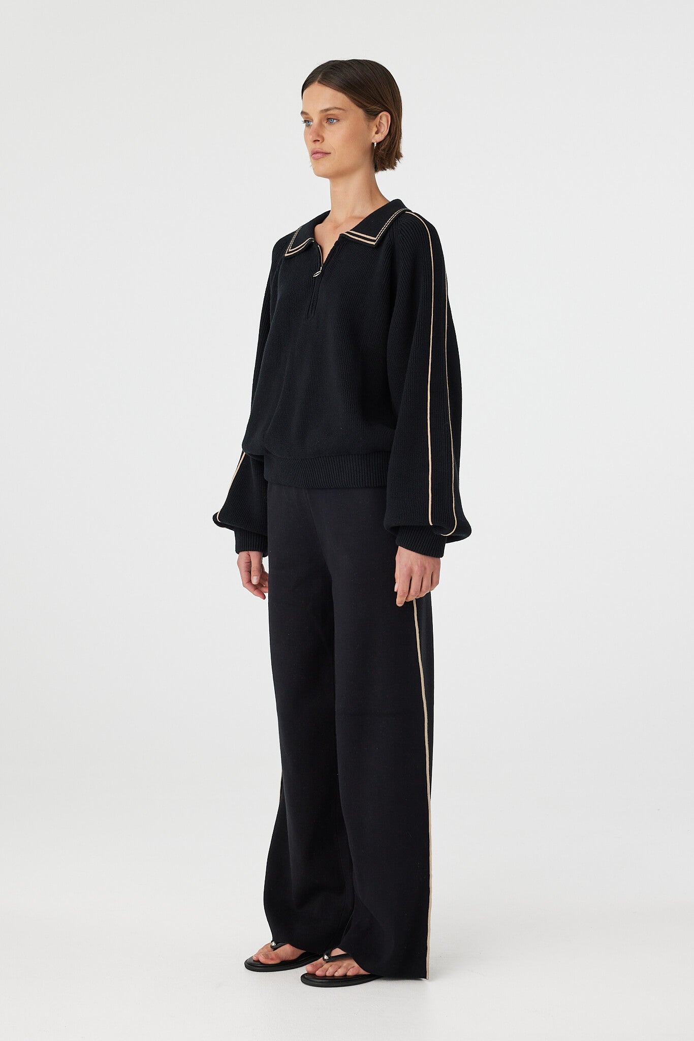 Nella zip sweater in black and taupe shown in side profile, highlighting the raglan sleeves, soft drape and contrast stripe detail along the arms, styled with black pants and sandals
