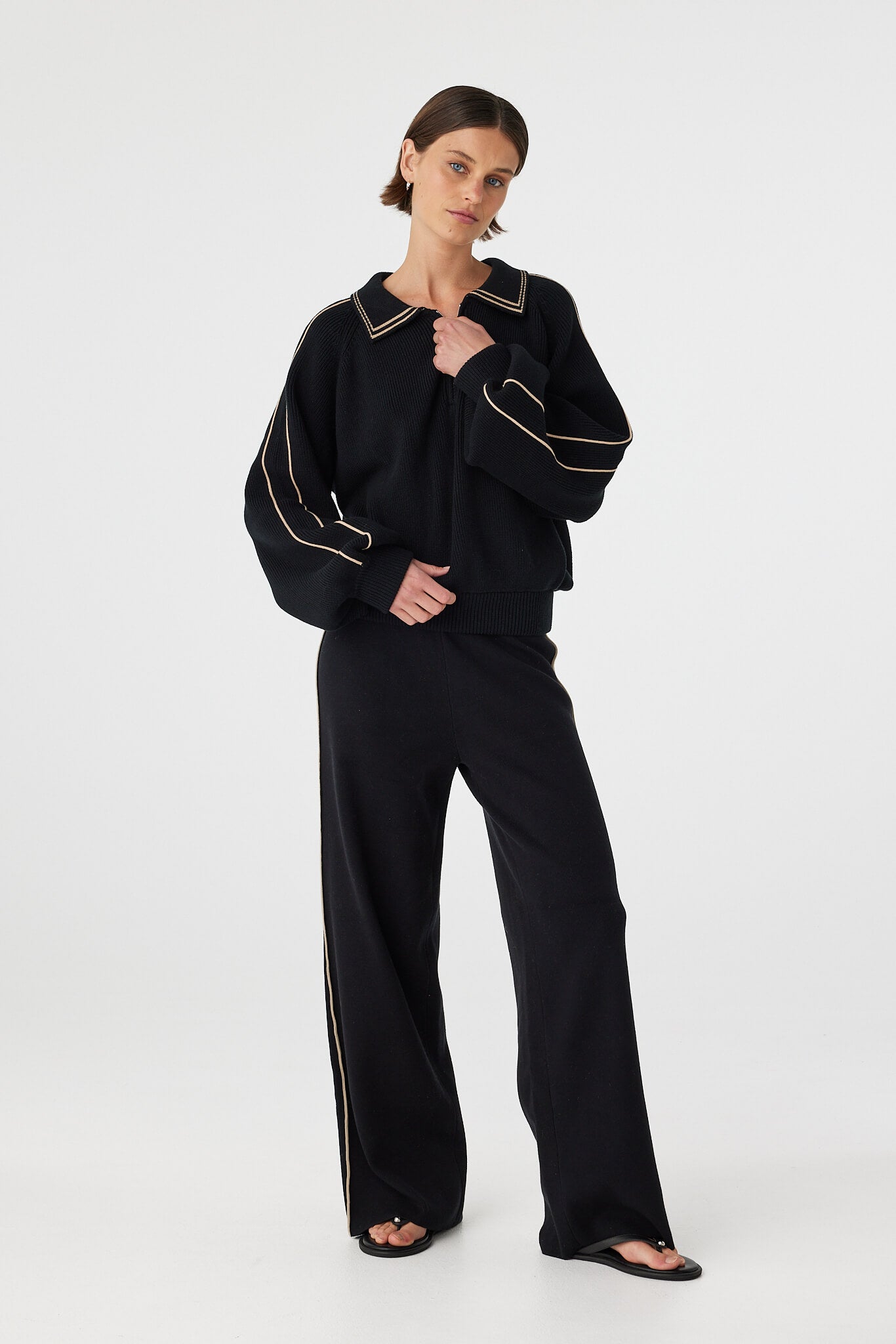 Nella zip sweater in black and taupe worn at an angle with hands near neckline, highlighting the zip closure, collar shape and sleeve volume, styled with black pants and sandals
