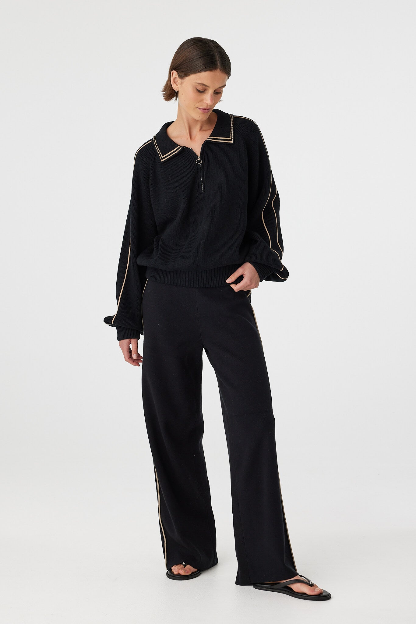 Nella zip sweater in black and taupe worn at an angle, showing the 1/4 metal zip, structured collar and relaxed fit, styled with black pants and sandals