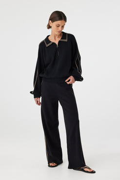 Nella zip sweater in black and taupe worn at an angle, showing the 1/4 metal zip, structured collar and relaxed fit, styled with black pants and sandals