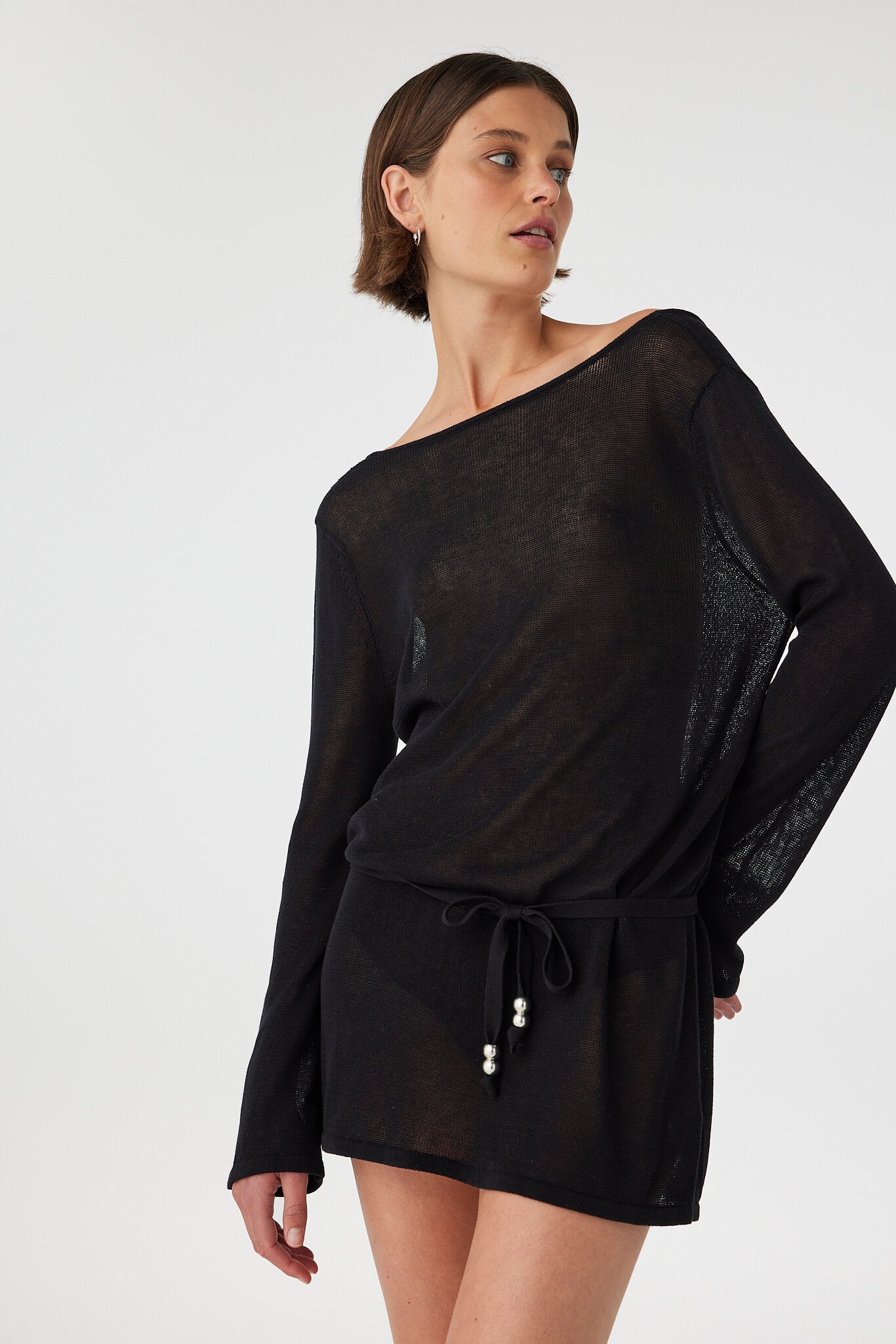 Sheer black cotton mini dress with long sleeves and removable waist tie, styled loosely to show adjustable shape and relaxed coastal layering.