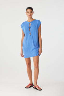 Model stands front-facing in pacific blue shift dress with twist detail worn at front, showing structured yet fluid fit in a breathable linen–cotton blend.