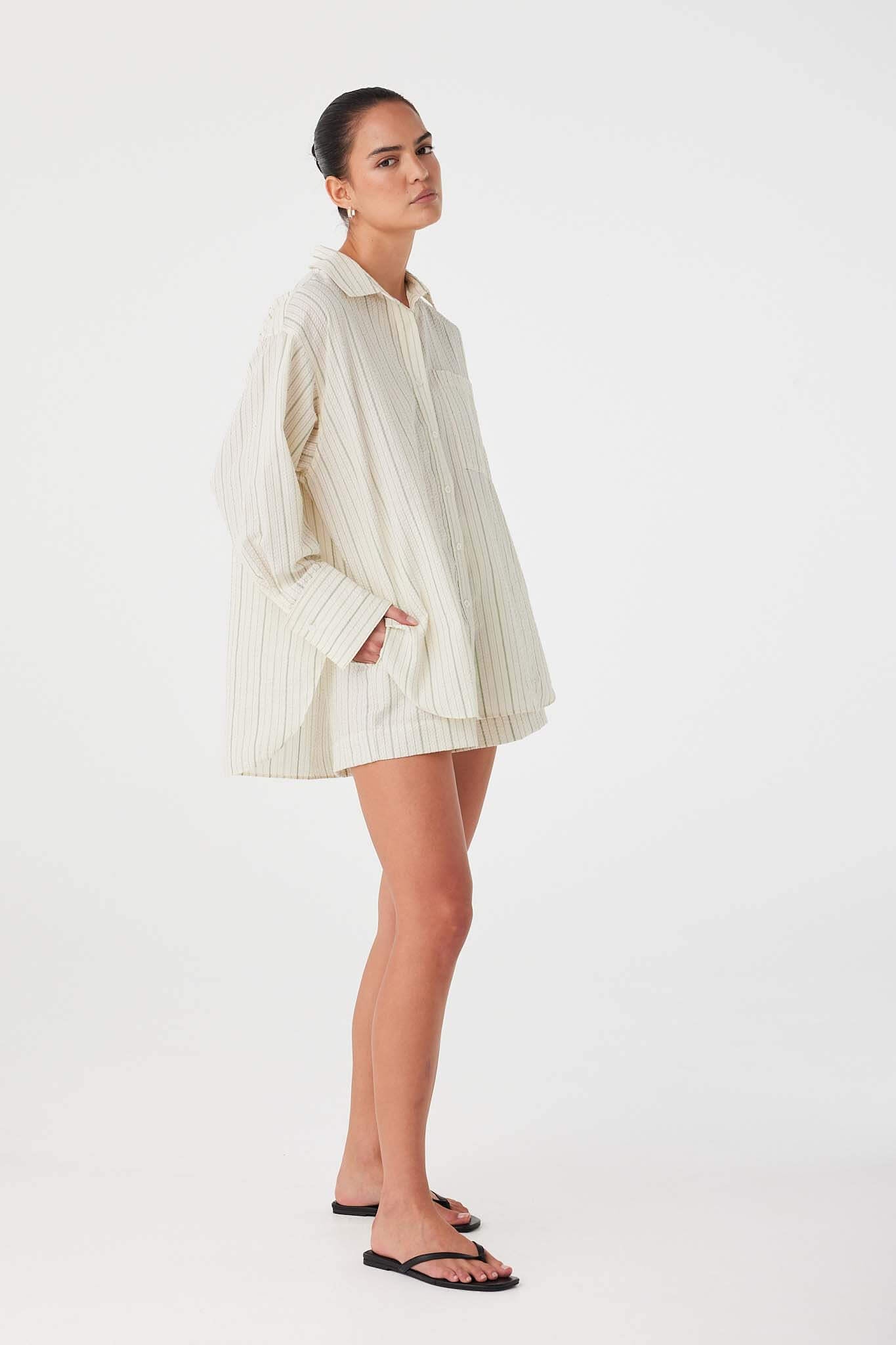 Model layering olive stripe cotton shirt in oversized fit, styled with matching shorts for coordinated set.