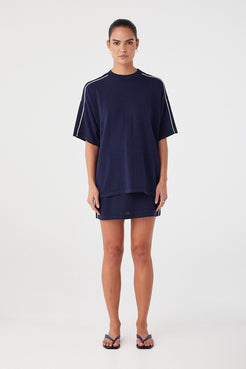 Model wears navy mini skirt in linen–cotton knit with contrast white side stripes. Straight silhouette with ribbed waistband and drawcord.
