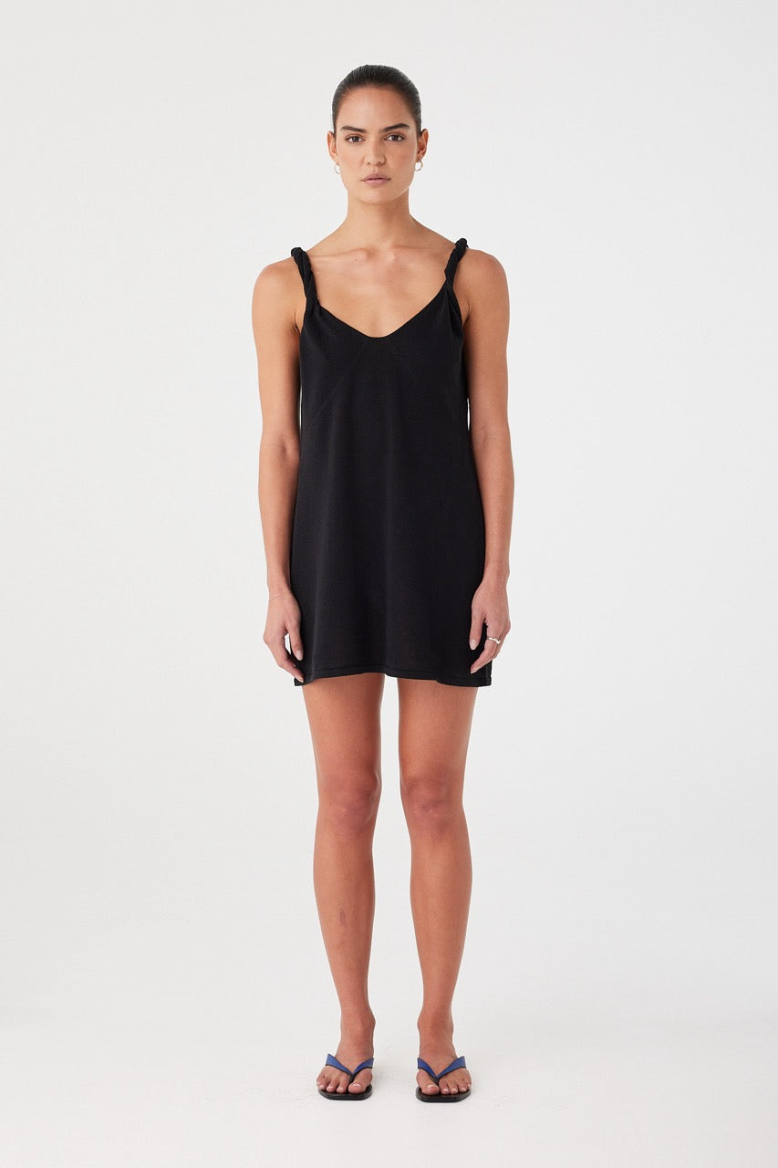 Model wearing black mini dress in linen–organic cotton blend. V neckline and twist straps with a semi-fitted silhouette for warm-weather ease.