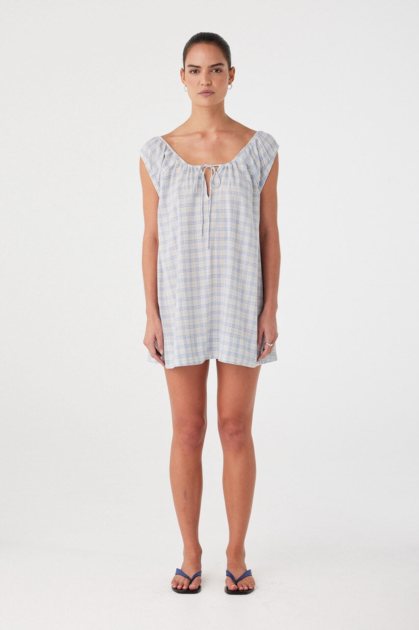 Model wears powder blue check mini dress with adjustable drawstring neckline and front keyhole. Flared silhouette in lightweight cotton.