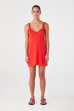 Model wears hibiscus red mini dress in linen–cotton blend. V neckline with twist straps and bust seam shaping.