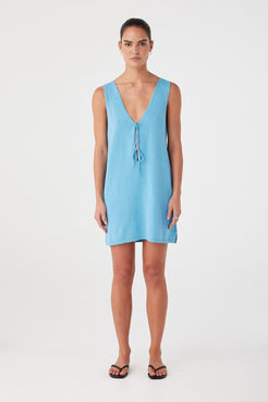 Model wears relaxed-fit mini dress in cyan linen-cotton with a V neckline and tie detail, styled for warm weather layering.