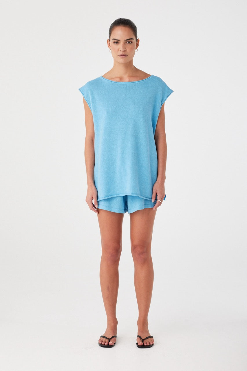 Model wearing oversized sleeveless top in cyan linen-cotton with side ties and rolled edges, styled for summer ease.