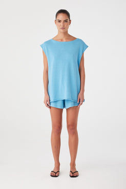 Model wearing oversized sleeveless top in cyan linen-cotton with side ties and rolled edges, styled for summer ease.