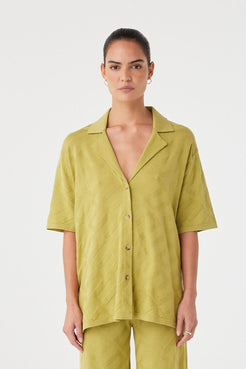 Model wears an oversized short-sleeve shirt in matcha green organic cotton knit. Breathable texture with button front and open collar.