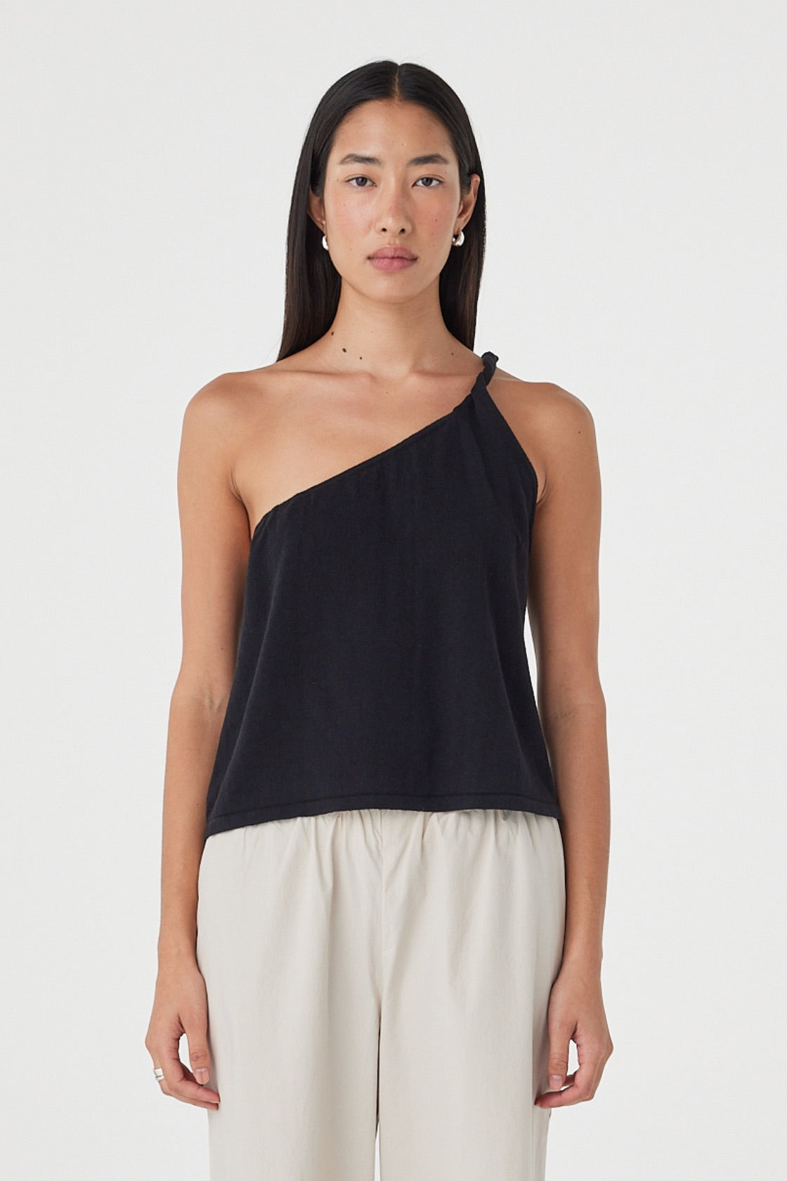 Front view of the Harri Asym Top in Black featuring an asymmetric neckline, one-shoulder twist detail and relaxed silhouette, styled with taupe pants.