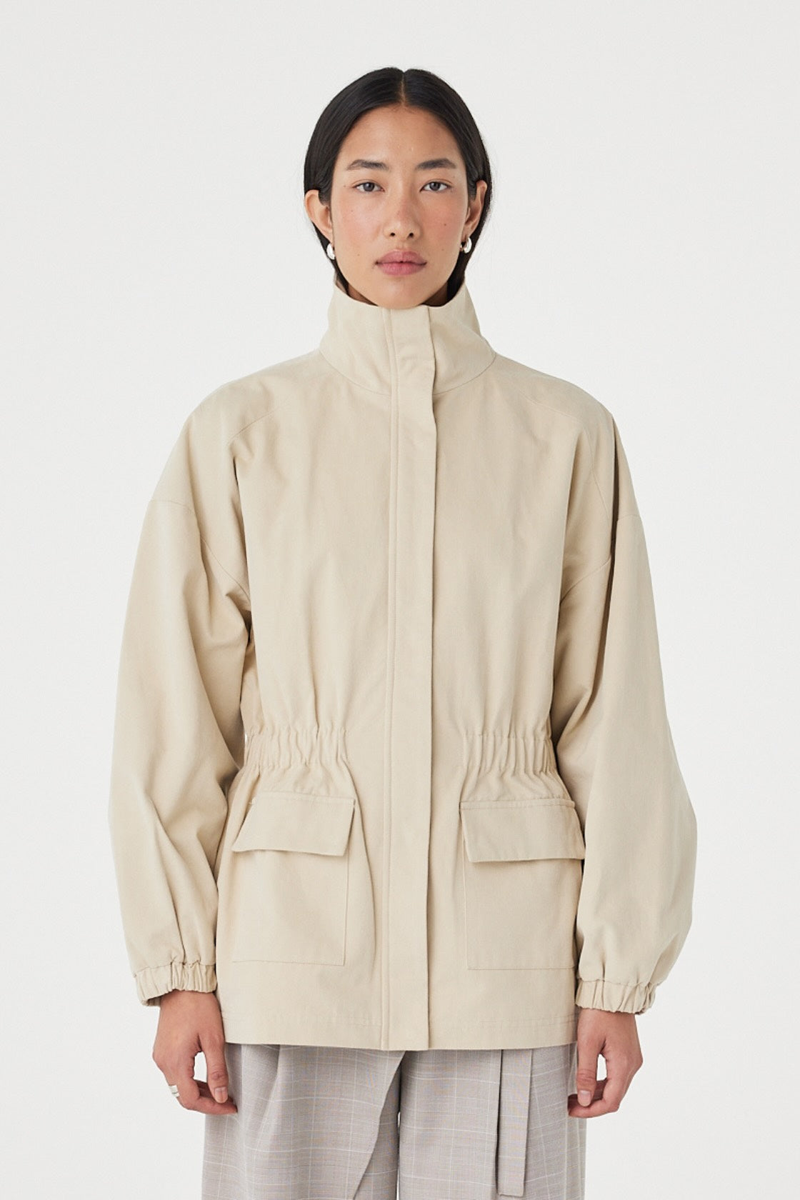 Front view of the Amari Jacket in Beige featuring a high neckline, concealed zip closure and gathered waist detail, styled with relaxed taupe trousers.