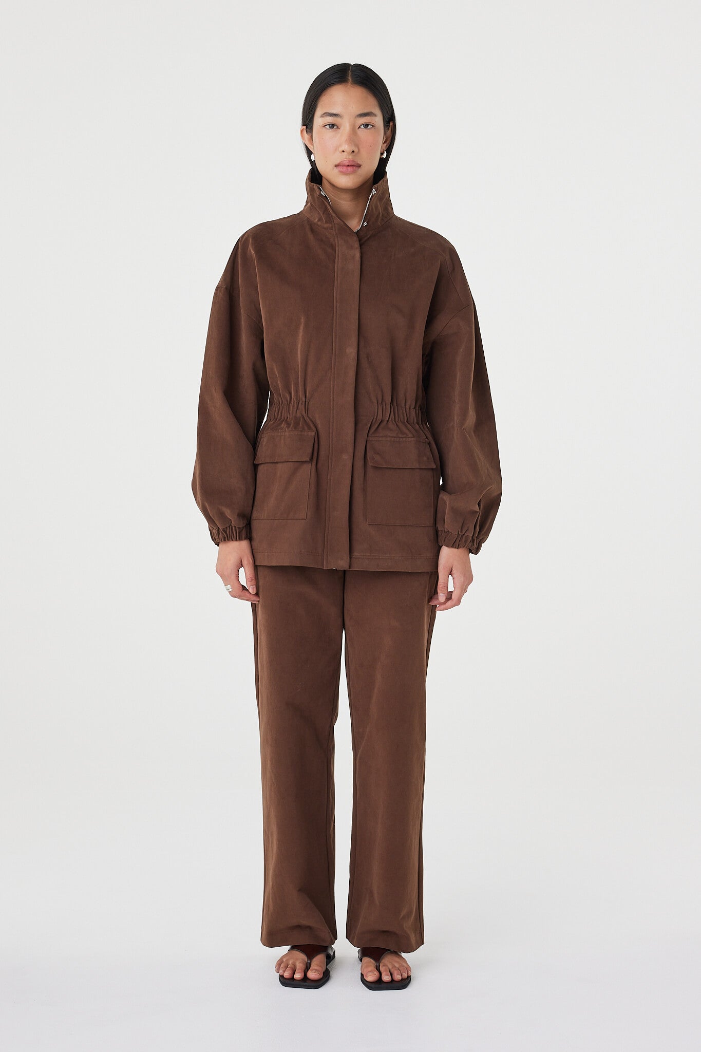 Amari Jacket in espresso with a soft velvet finish, styled with matching straight-leg pants and black sandals, featuring oversized flap pockets and a high funnel neckline