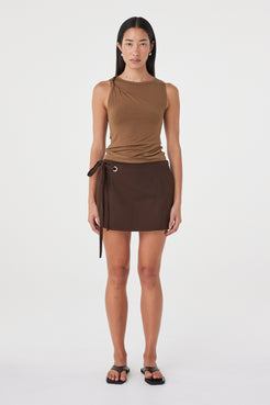 Front view of the Lucia Tie Mini Skirt in Espresso featuring an asymmetrical wrap panel, silver eyelet and double tie detail, styled with a brown tank.
