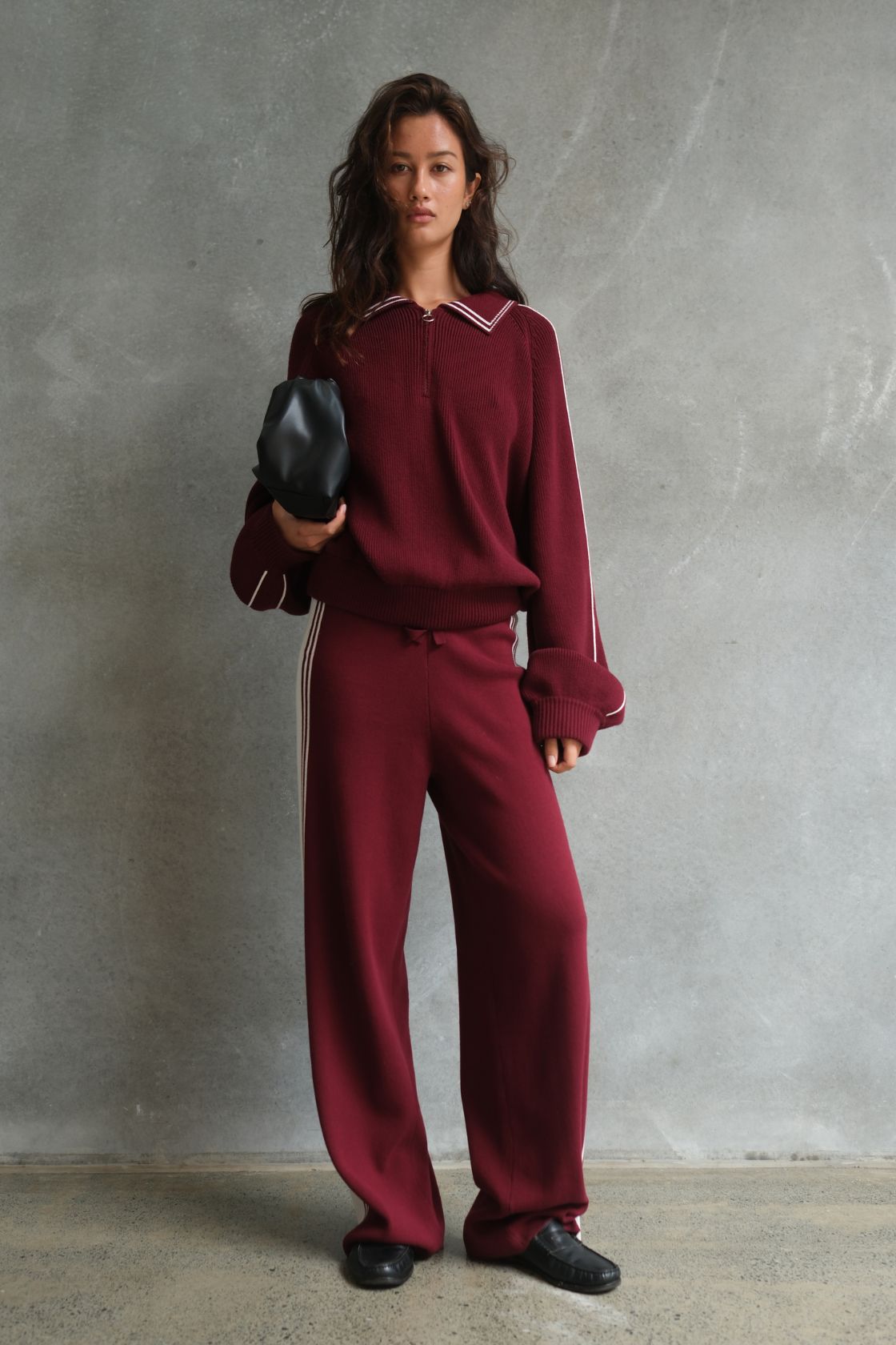 Model wearing the Emery Pant in Merlot & Cream, featuring a relaxed wide-leg silhouette with contrast cream side stripes, styled with the matching Nella Zip knit and a black pouch bag against a textured concrete backdrop.