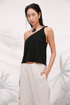 A woman modeling a black one-shoulder crop top with white wide-leg pants, standing against a white backdrop with soft tropical leaf patterns.